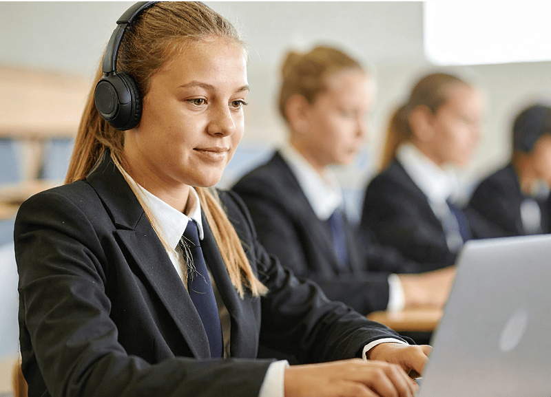 Students in school assembly hall wearing school uniforms completing exams on laptops.