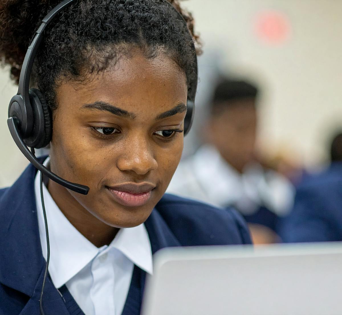Student wearing a headset and uniform, working on a laptop during exam. Other students can be seen blurred in the the background.
