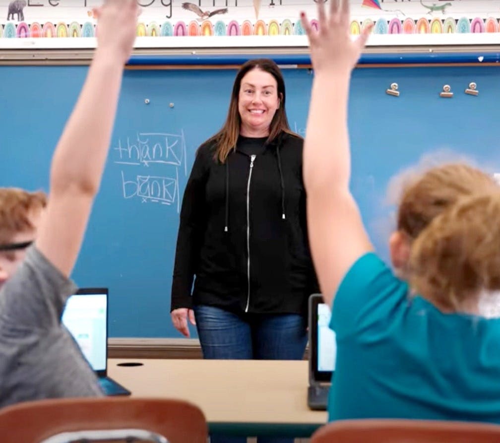 A teacher standing at the front of the class while two students raise their hands.