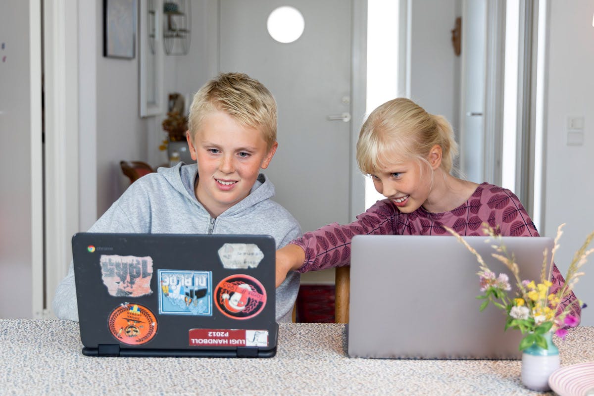 Två barn sitter vid ett bord med varsin laptop, medan flickan pekar mot pojkens skärm och de båda ler.