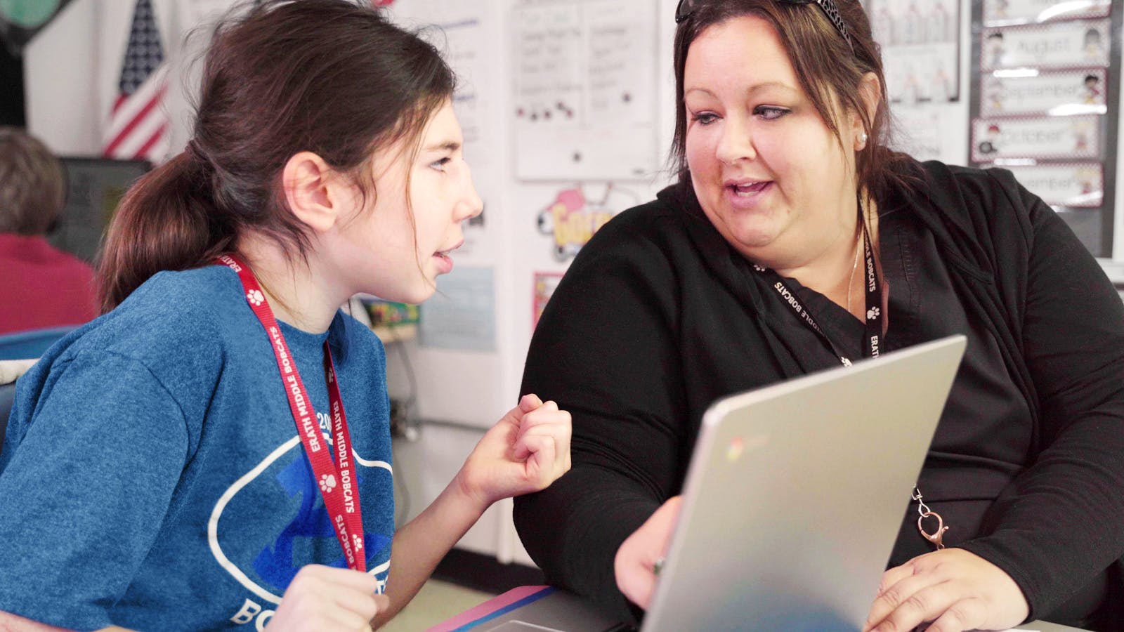 Student and teacher sit together at a desk, talking to each other while working on a laptop.