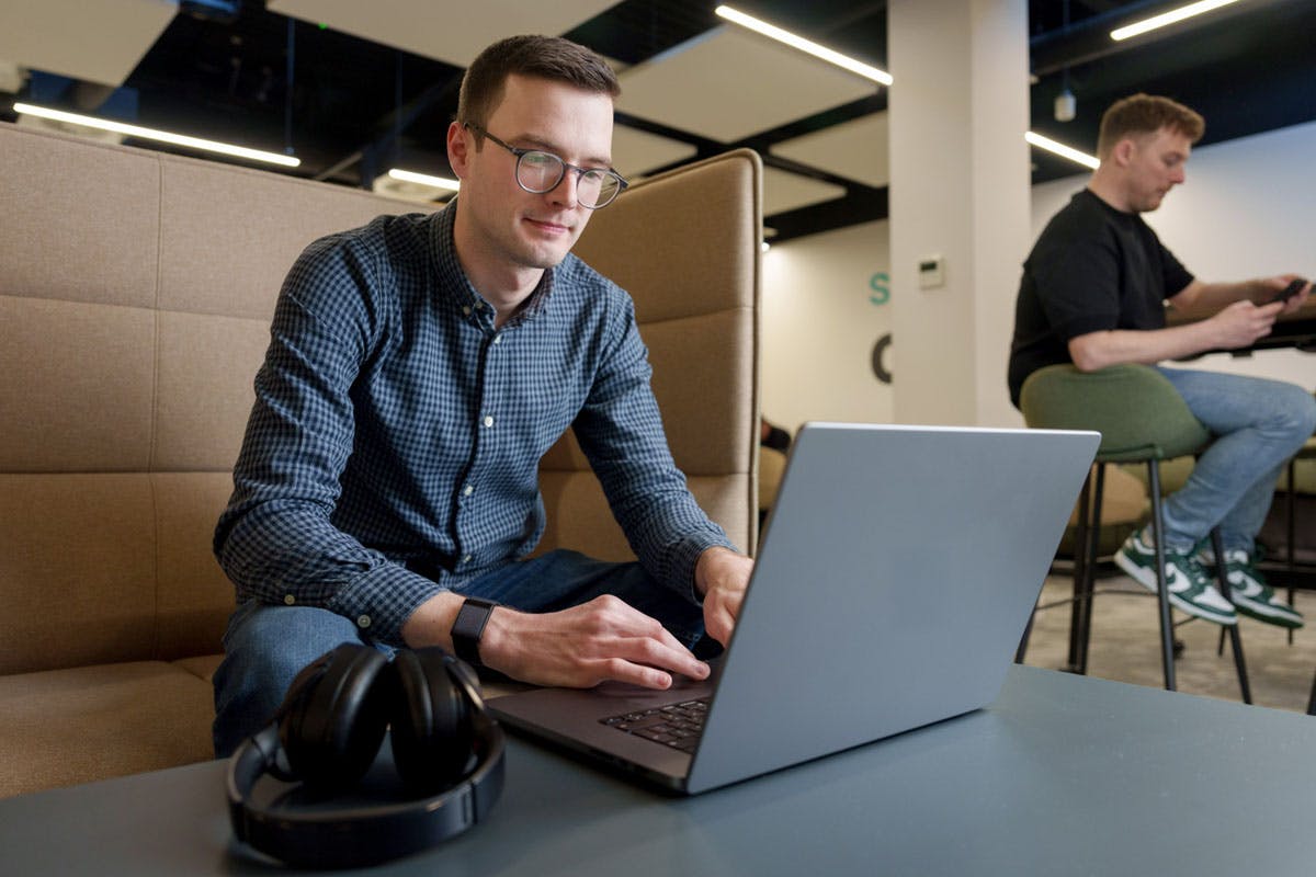 Mand arbejder på en bærbar computer ved et bord i et kontormiljø, med høretelefoner foran sig og en kollega i baggrunden.