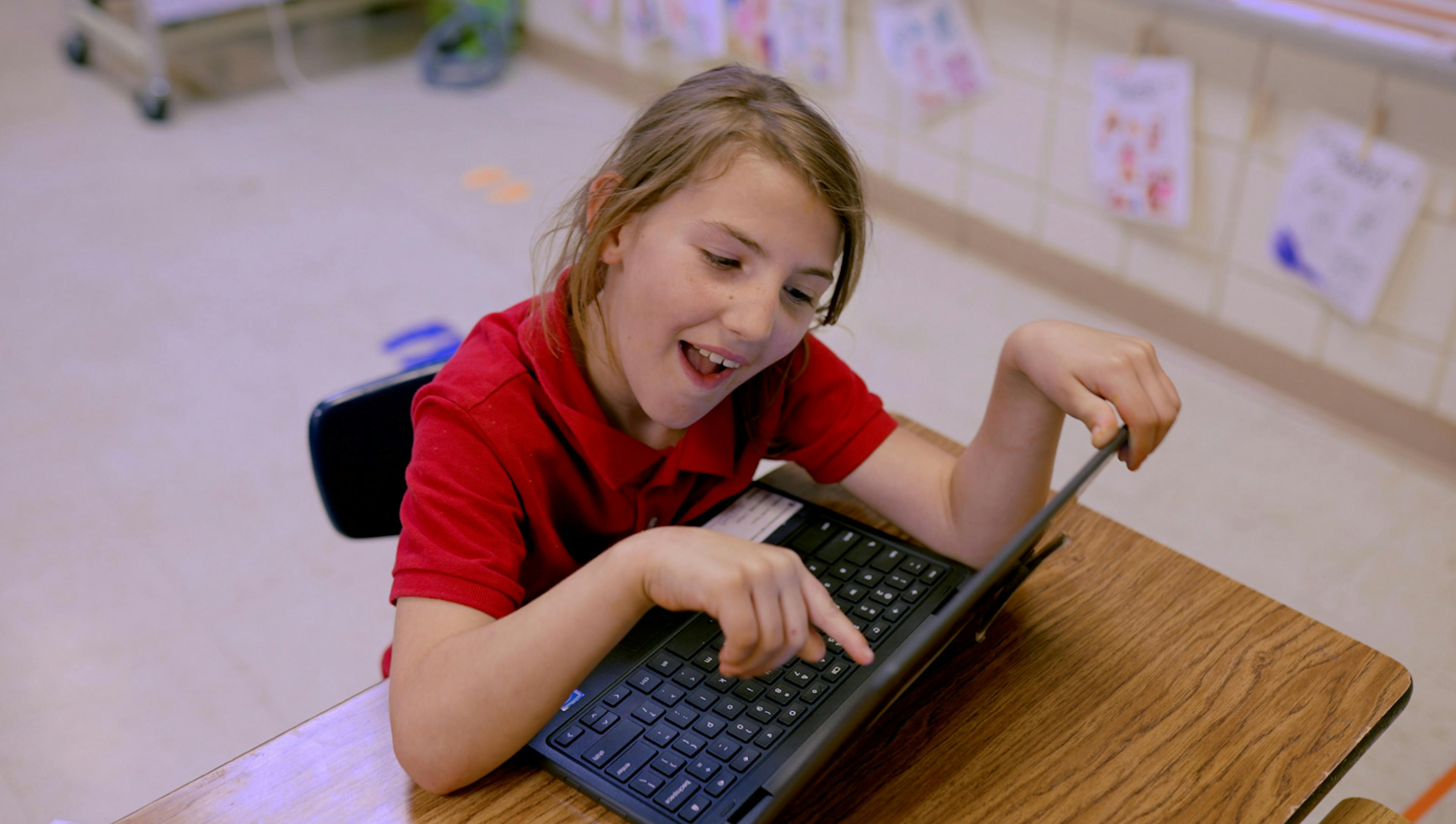 Child in a red shirt smiling while using a laptop at a classroom desk.