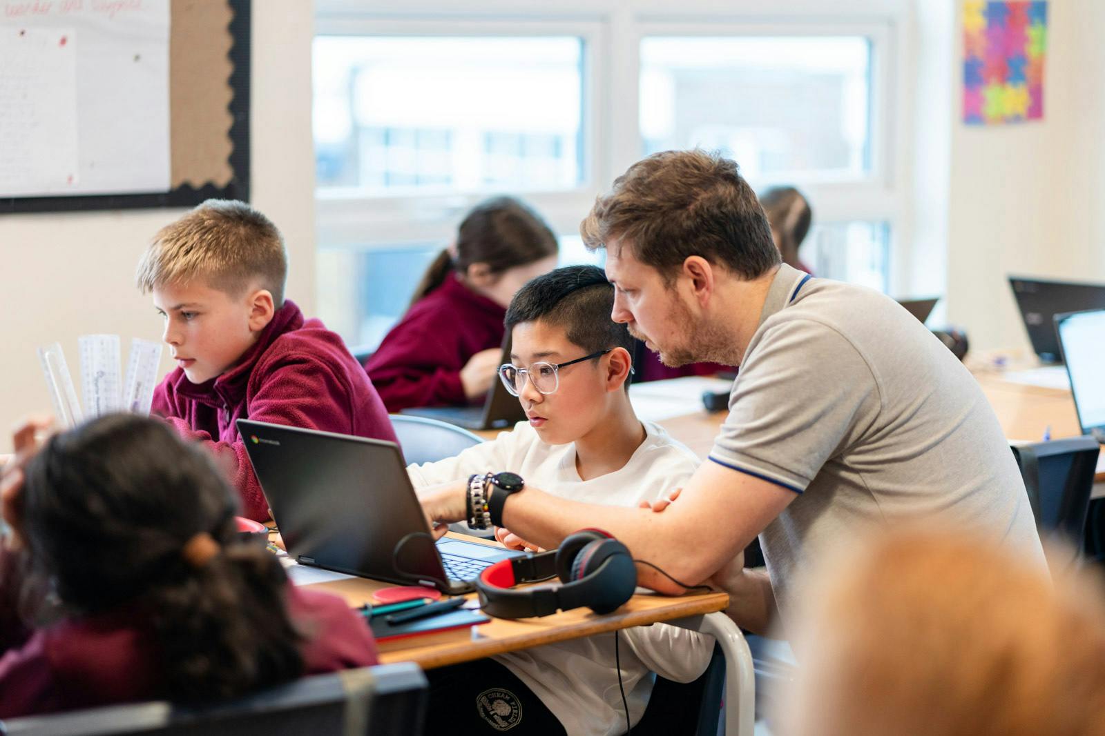 Teacher assisting a student with work on a laptop in a busy classroom.