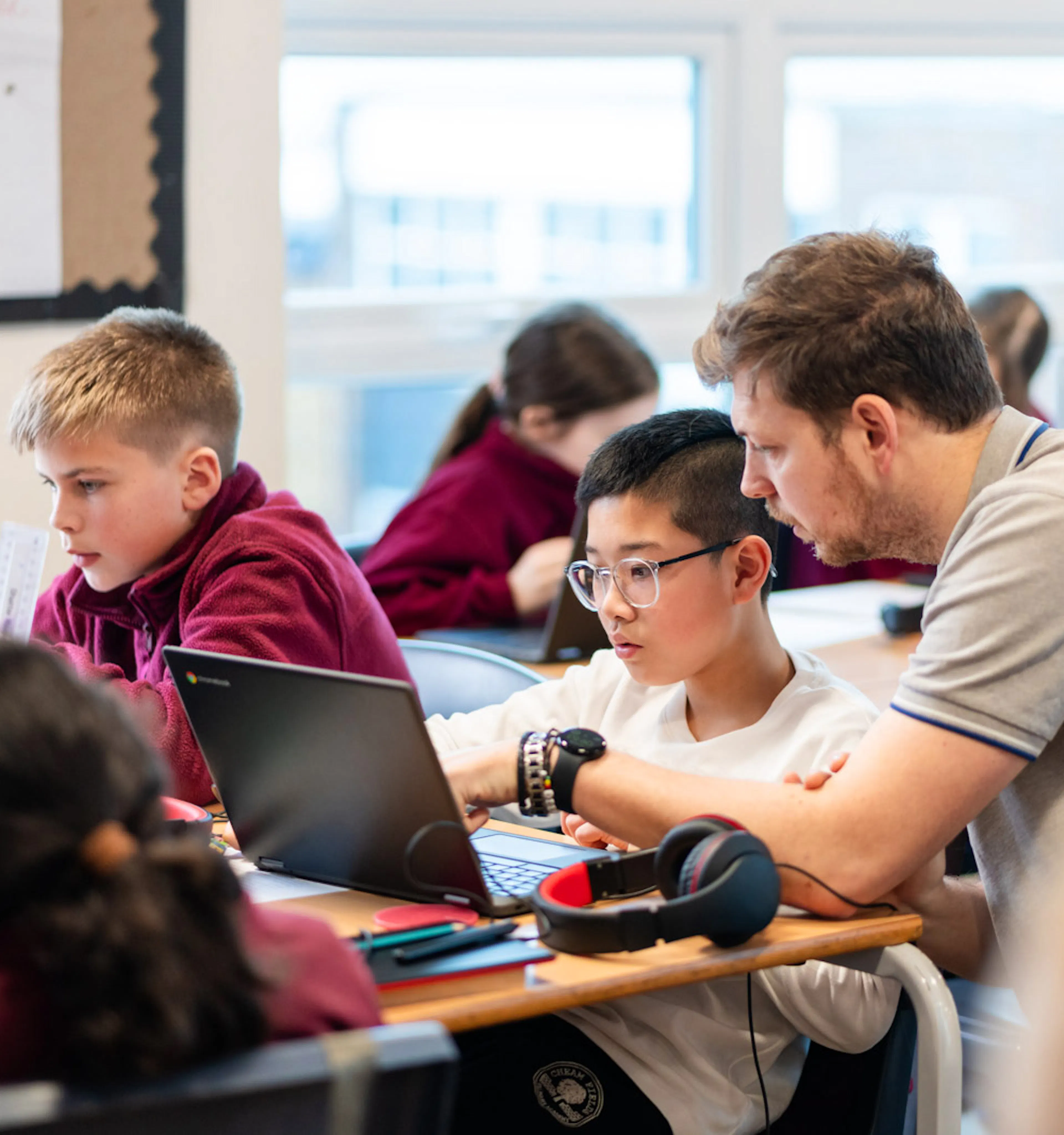 Teacher assisting a student with work on a laptop in a busy classroom.