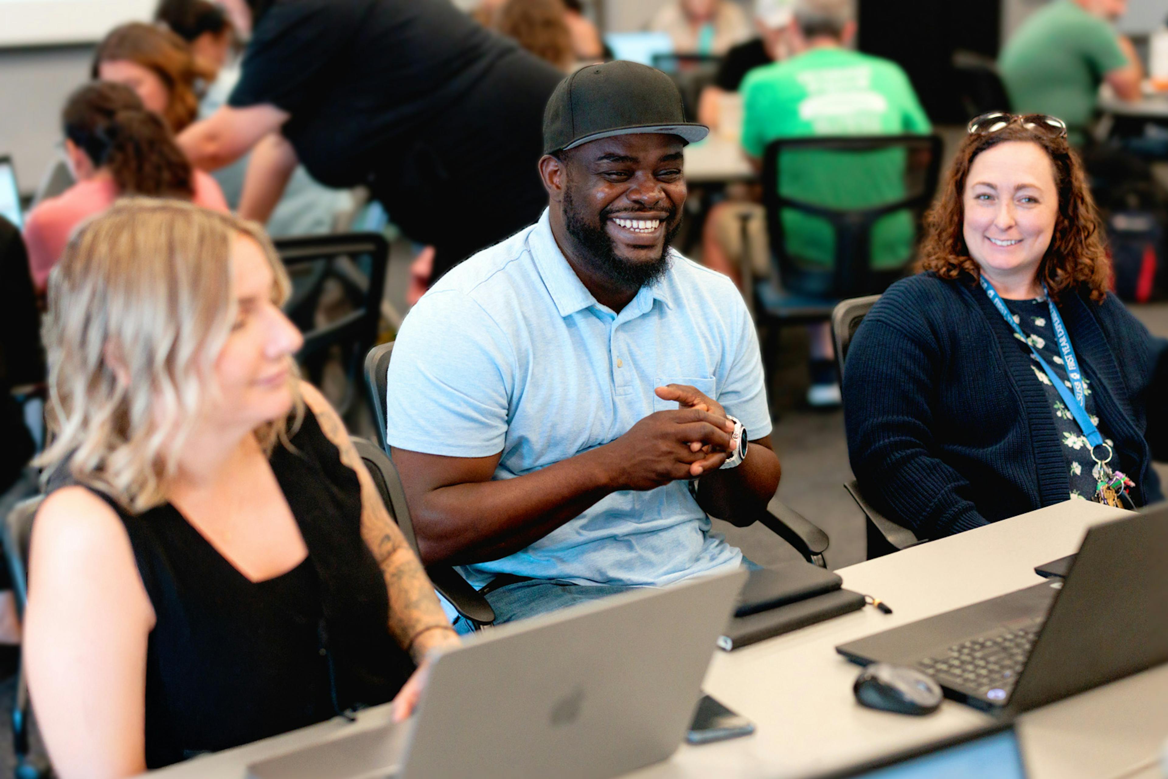 Three adults seated at a table with laptops, smiling and talking during a collaborative workshop or training session.