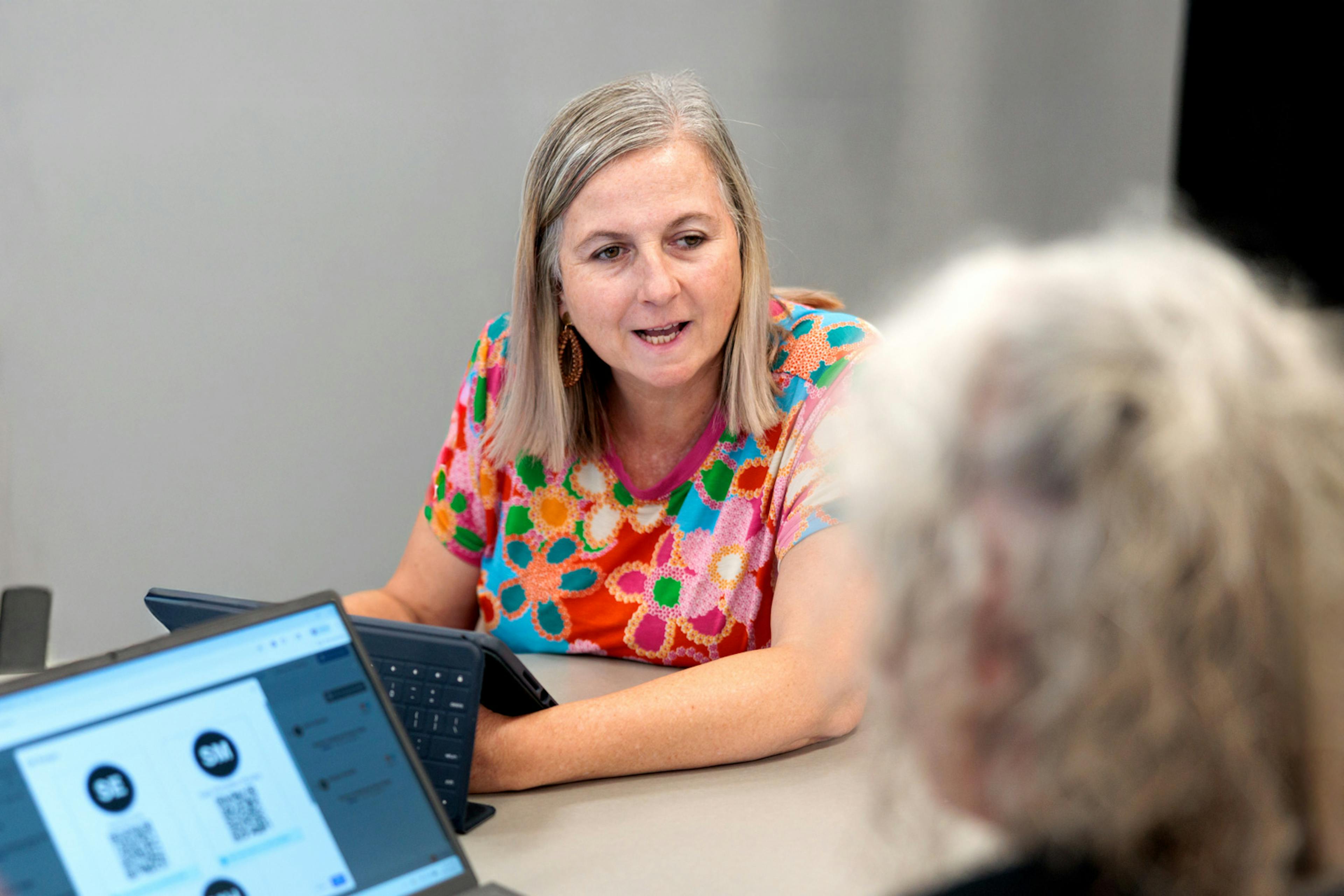 Woman speaking with a colleague across a table while using a tablet during a collaborative workshop.