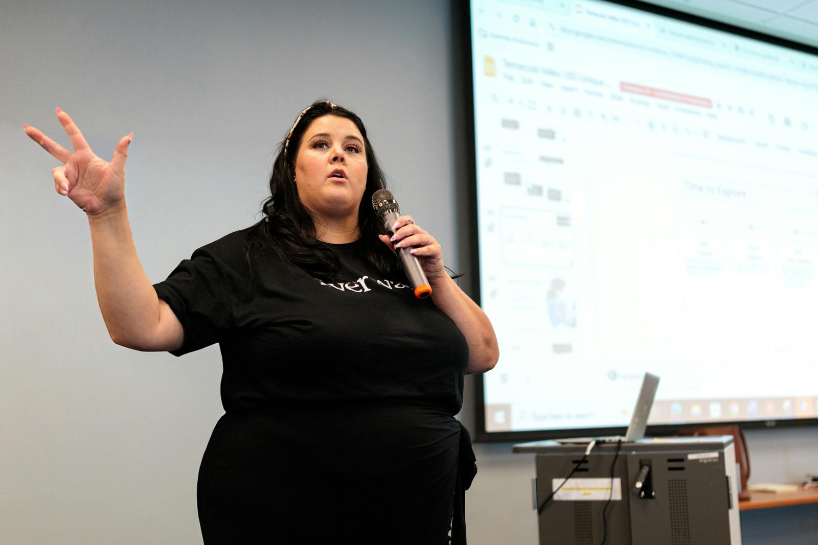 Woman holding a microphone and gesturing while presenting in front of a projected screen during a workshop or training session.