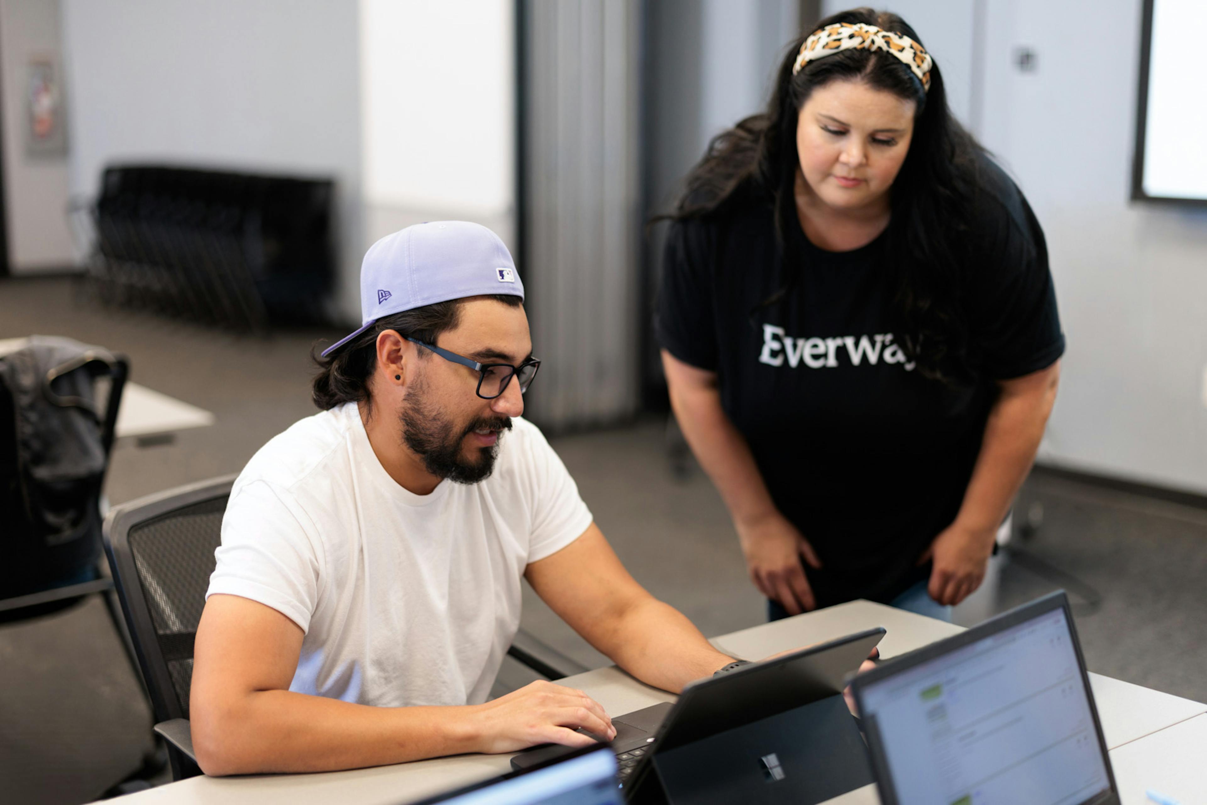 Man working on a laptop while a woman leans over to assist him during a classroom or training session.