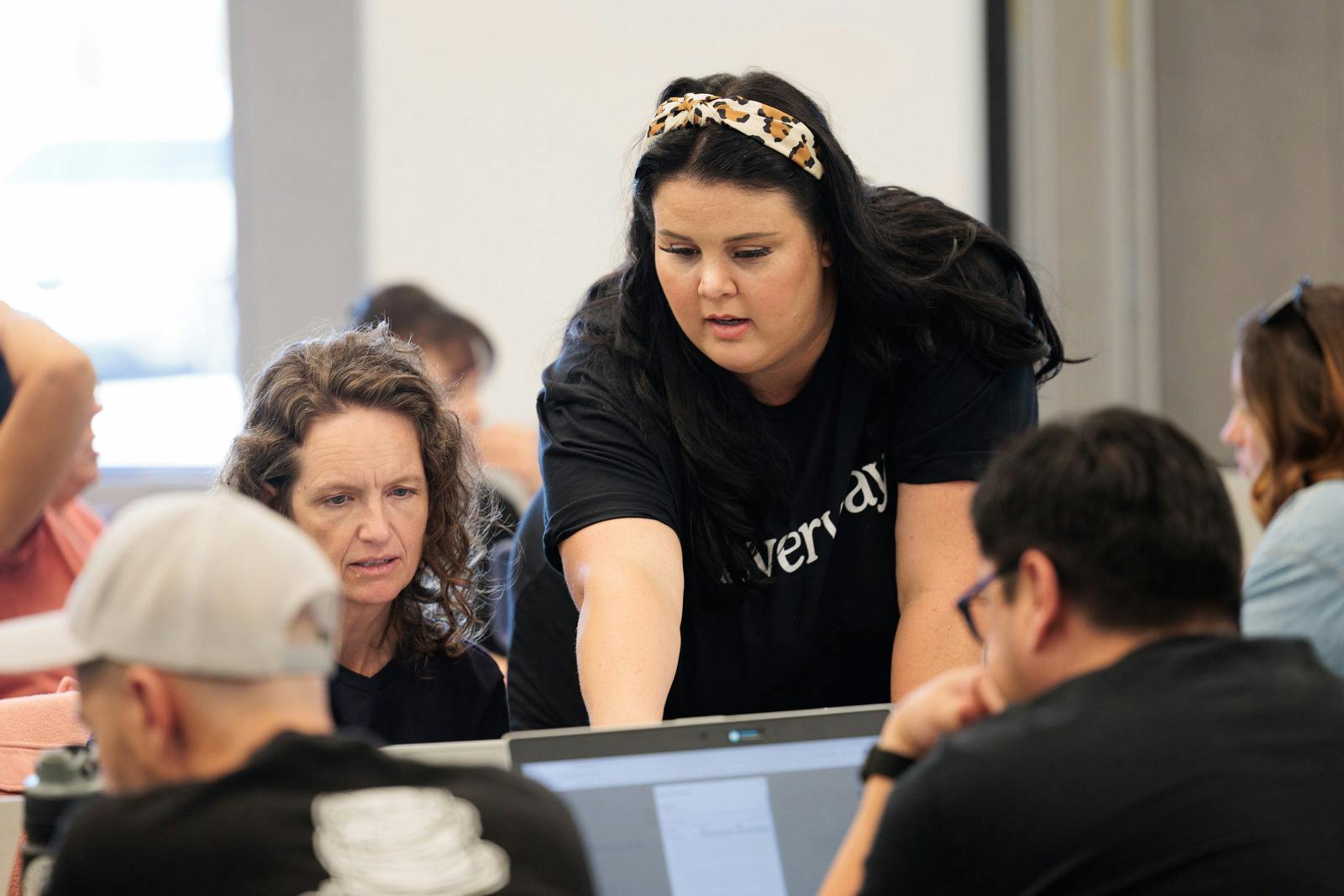 Woman assisting a group of adults during a computer-based workshop, pointing at a laptop screen to guide discussion.