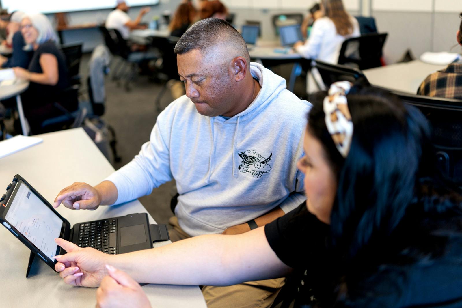 Two people sit at a table working together on a tablet, discussing something on the screen in a classroom setting.