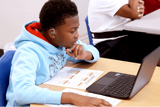 Student wearing a light blue hoodie working on a laptop with assignment sheets on the desk.