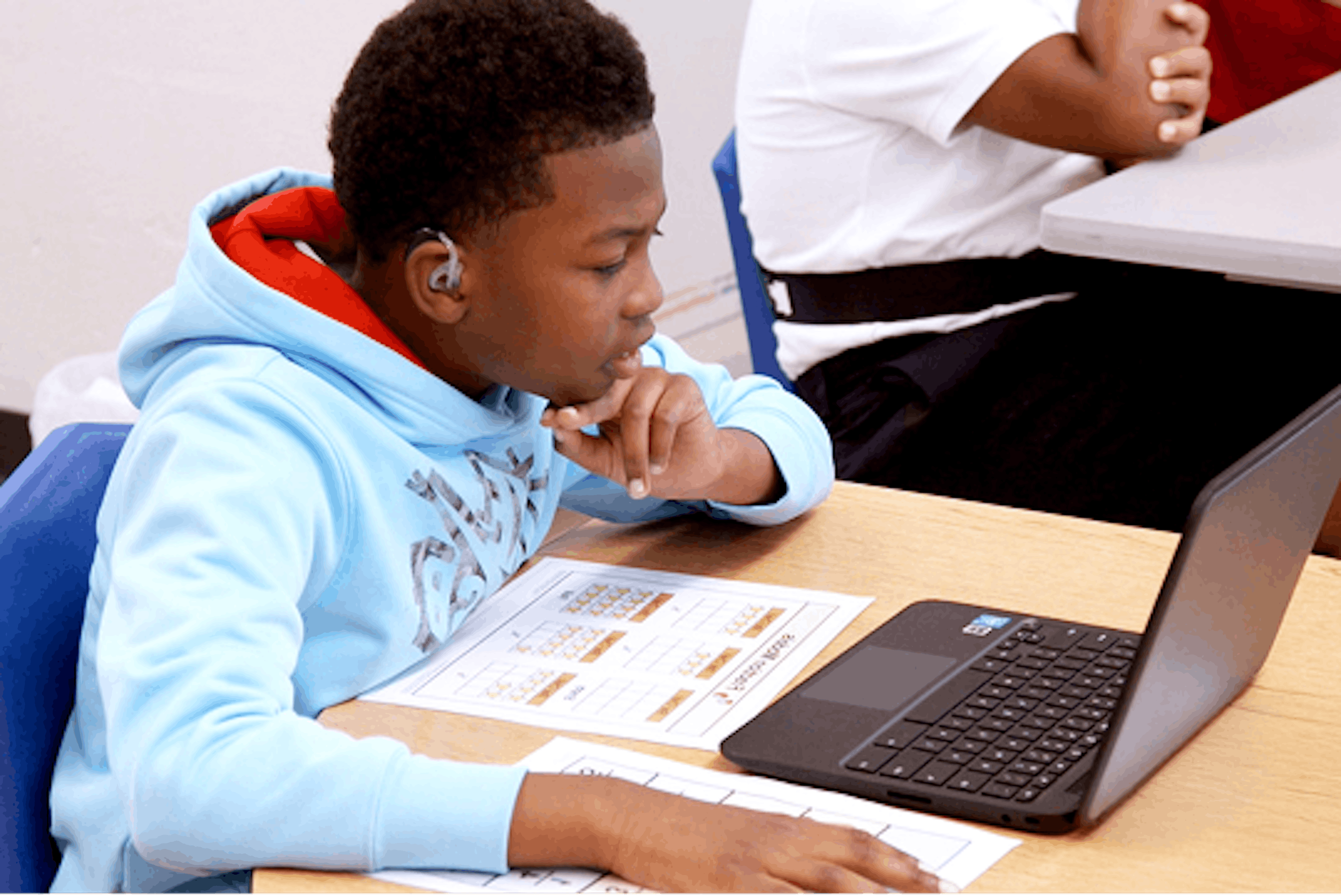 Student wearing a light blue hoodie working on a laptop with assignment sheets on the desk.