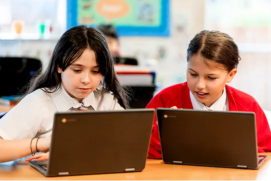 Two students using laptops together at a classroom desk, collaborating on a learning activity.