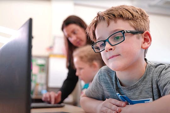 Young student wearing glasses focused on a laptop, with teacher assisting another child in the background.