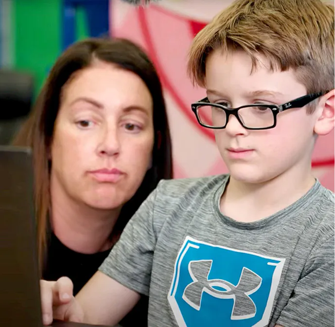 Teacher assisting a student wearing glasses as they work on a laptop together.