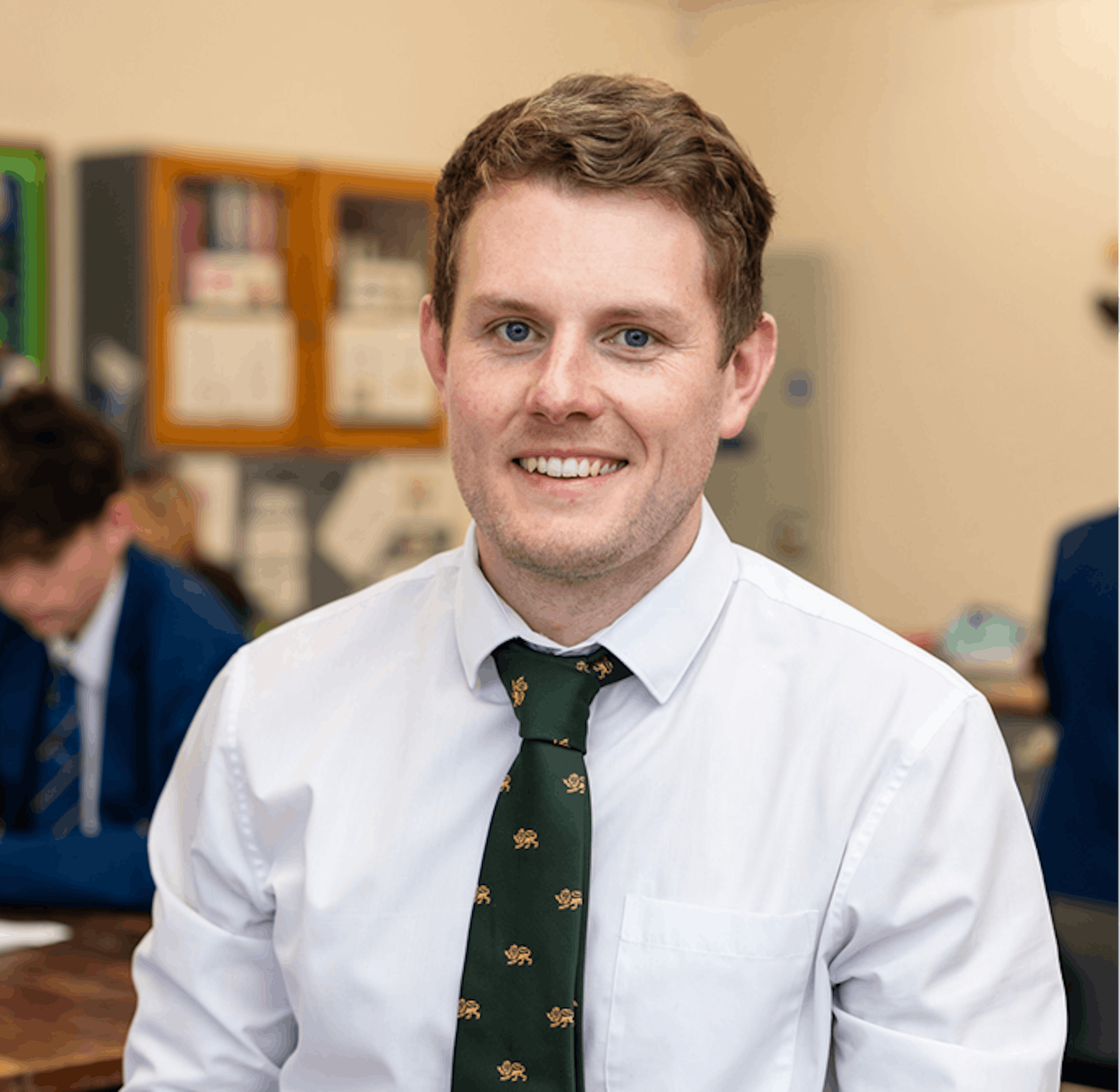 Teacher wearing a white shirt and green tie smiling in a classroom with students working in the background.