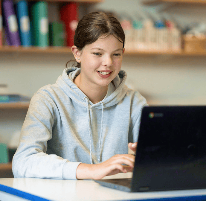 Student wearing a gray hoodie using a laptop at a desk in a classroom setting.
