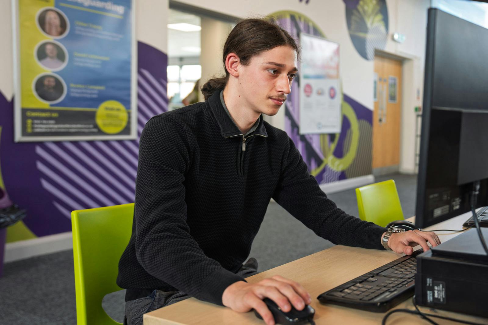 Student sitting at a desk using a desktop computer in a modern classroom or study area with colourful wall posters in the background.