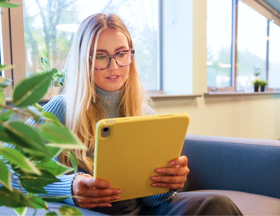Woman sitting on a couch working from a yellow tablet in a bright office with large windows and plants.