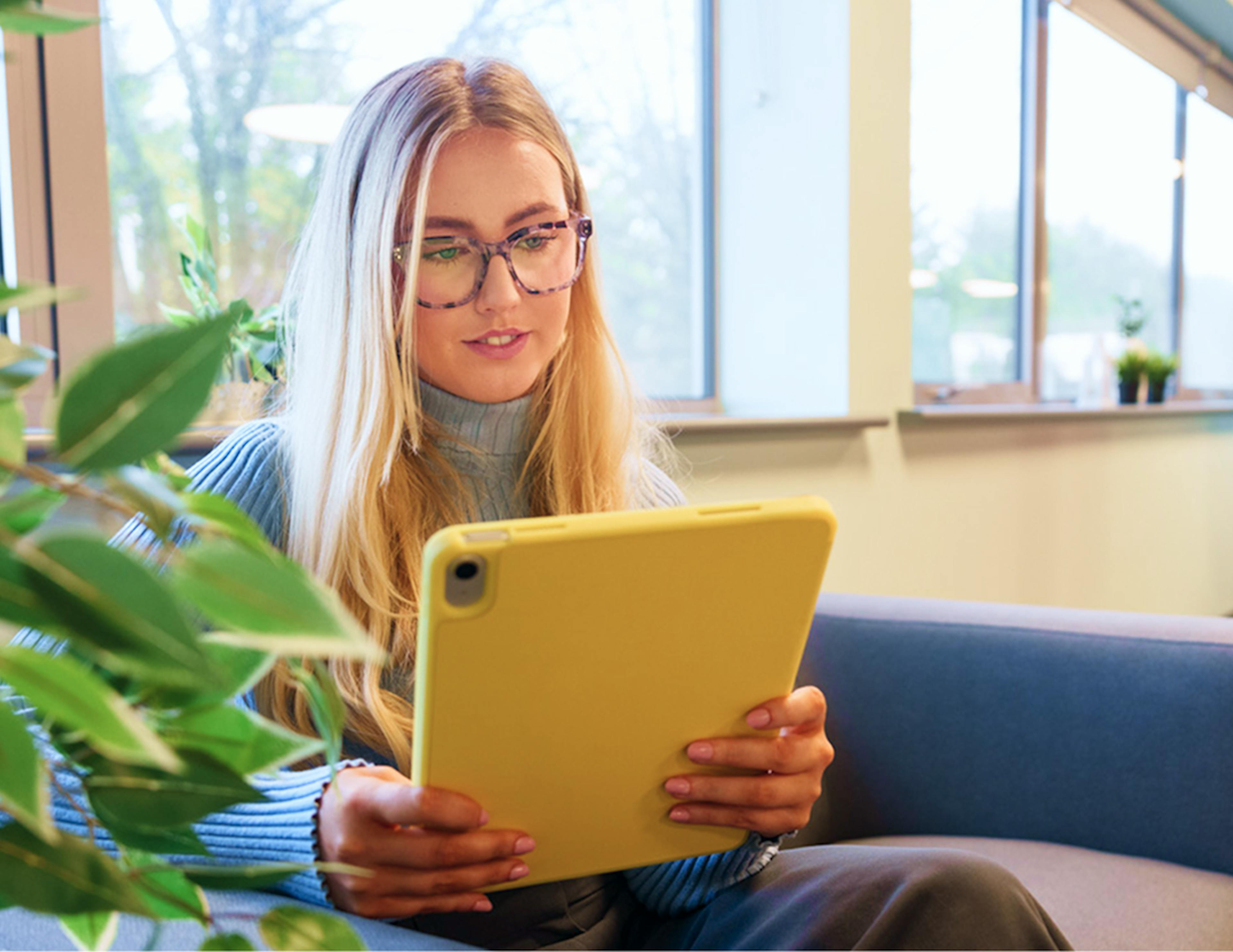 Woman sitting on a couch working from a yellow tablet in a bright office with large windows and plants.