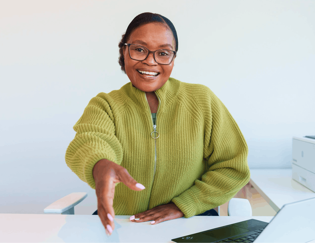 Smiling woman in a green sweater extending her hand for a handshake while sitting at a desk with a laptop.
