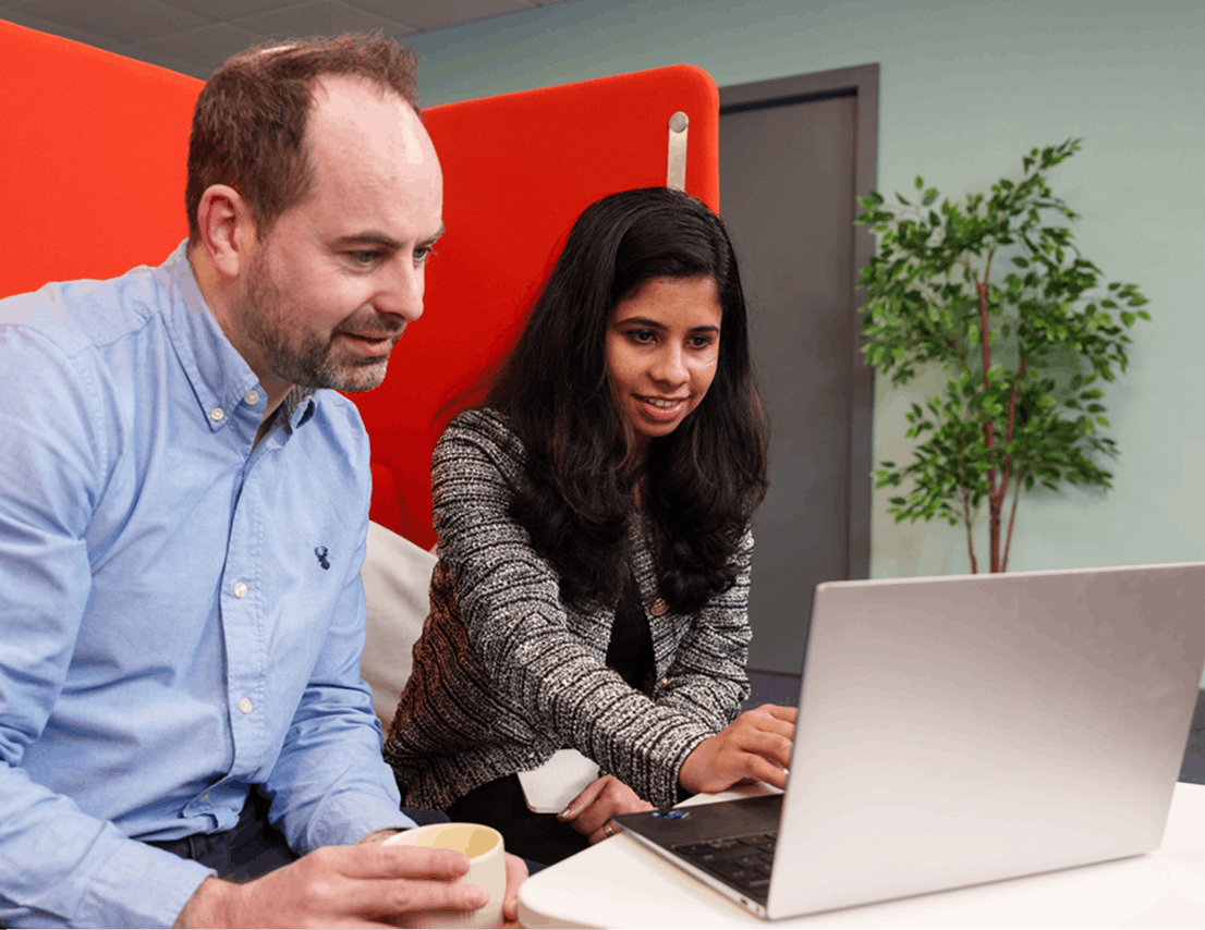 Two colleagues sitting together looking at a laptop screen, one holding a coffee mug while the other types.