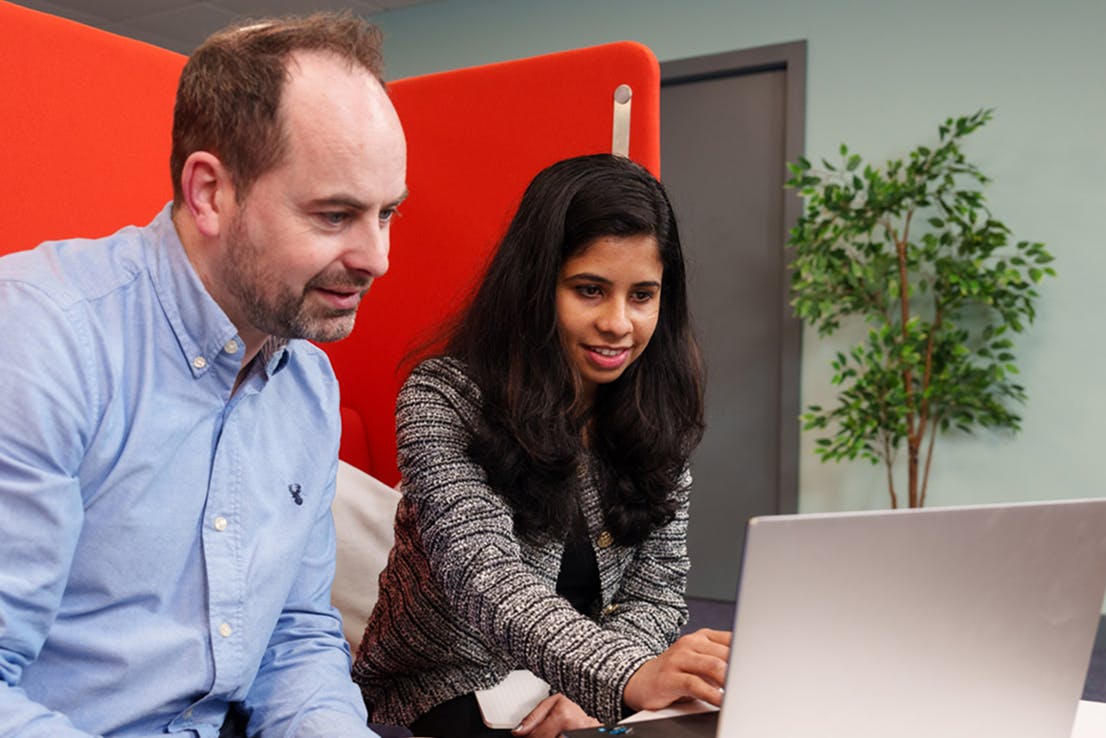 Two colleagues sitting together looking at a laptop screen, one holding a coffee mug while the other types.