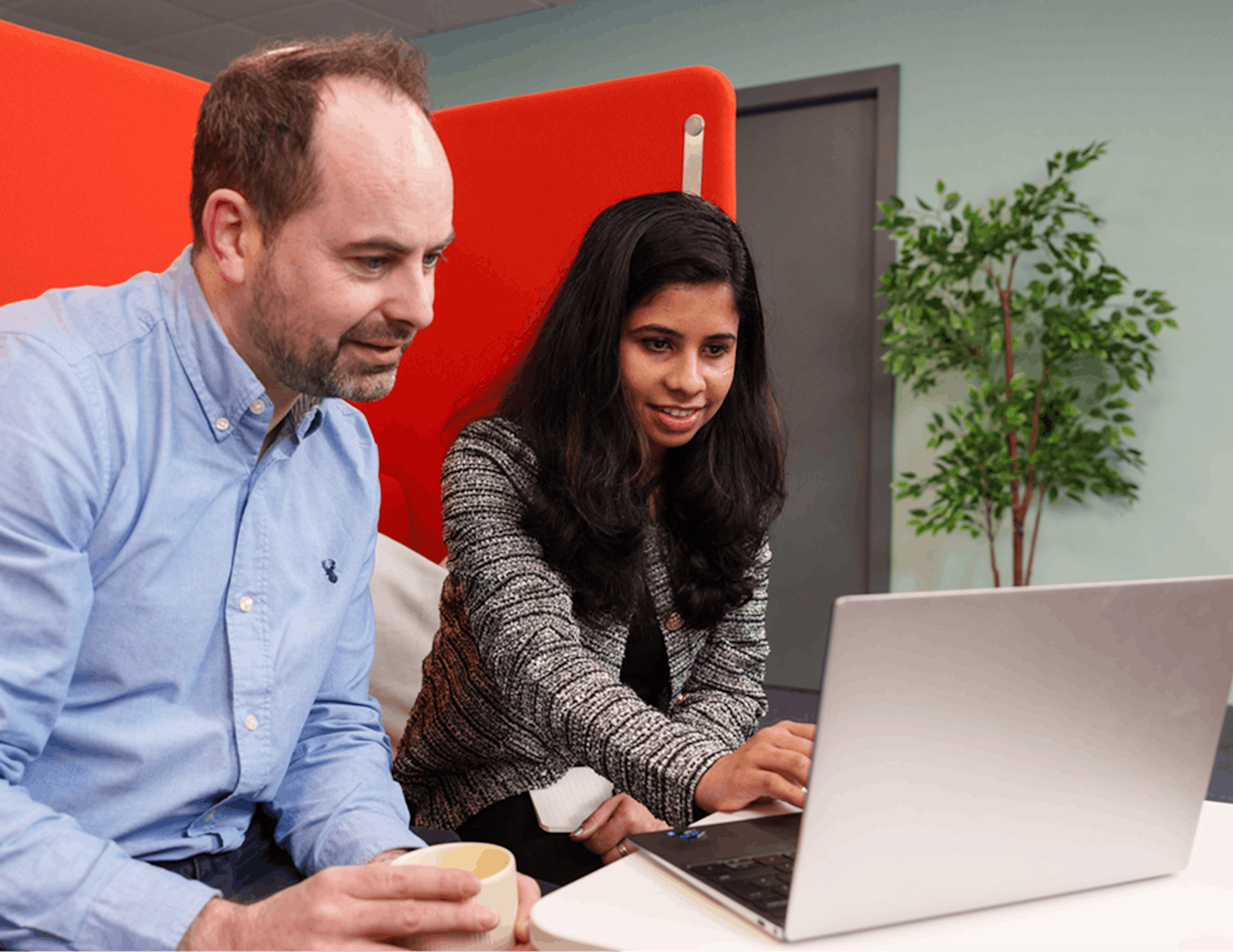 Two colleagues sitting together looking at a laptop screen, one holding a coffee mug while the other types.