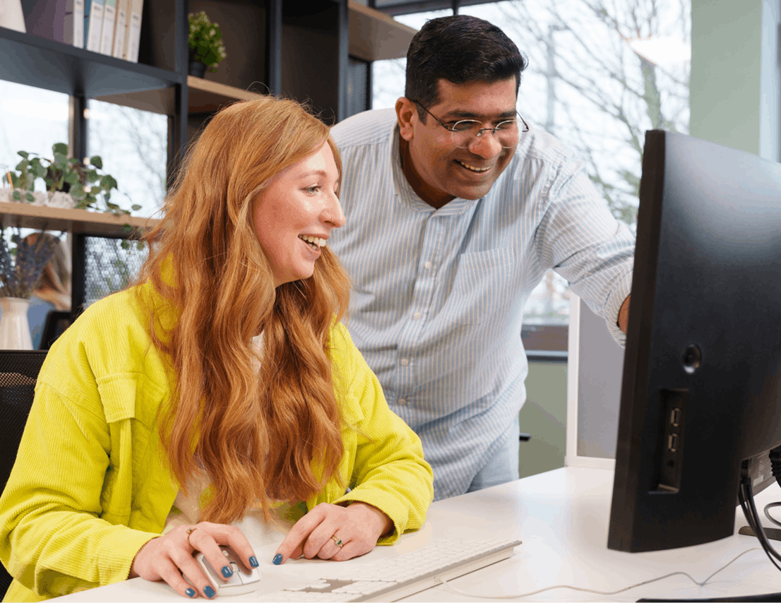 Two colleagues smiling and collaborating at a computer in an office workspace.