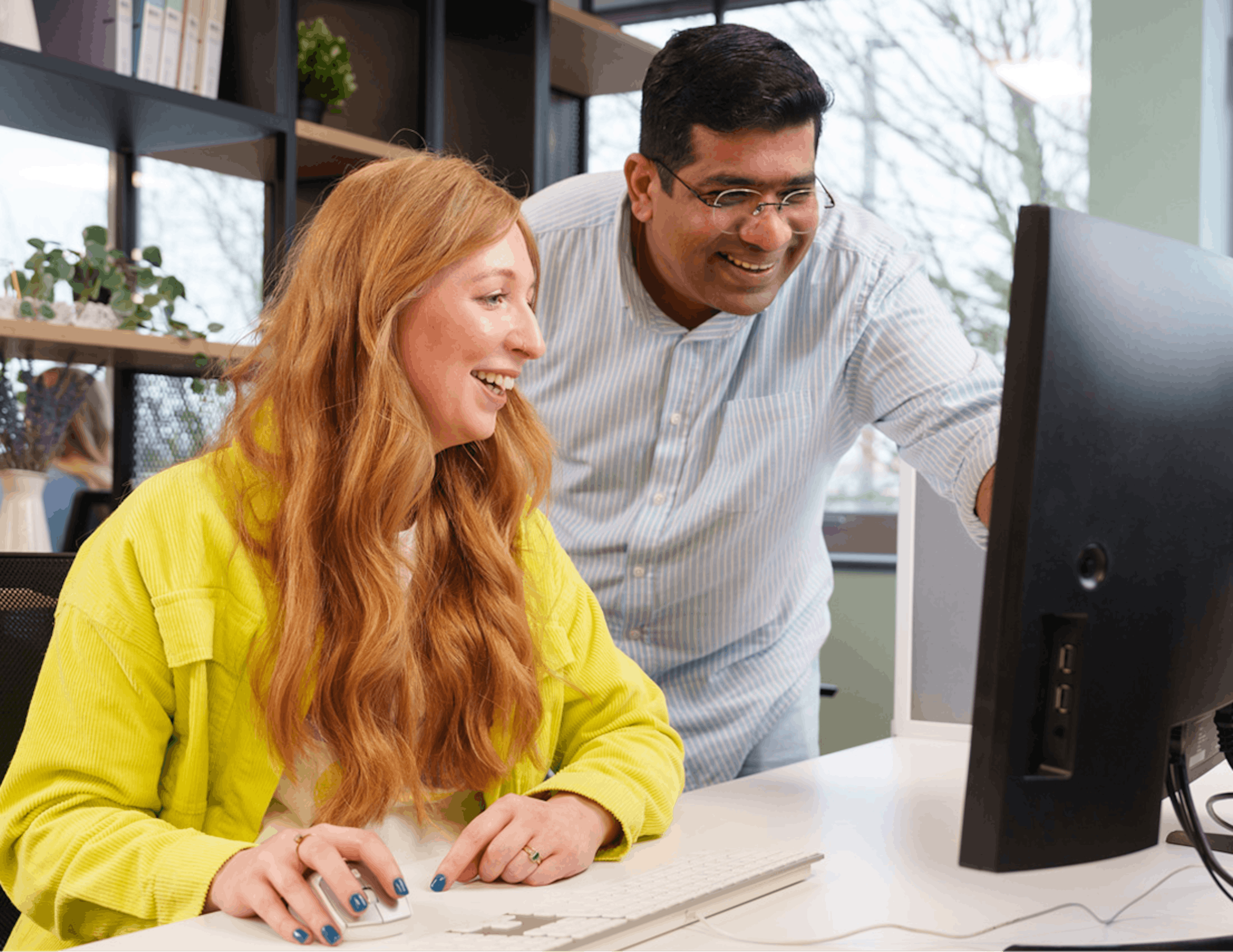 Two colleagues smiling and collaborating at a computer in an office workspace.