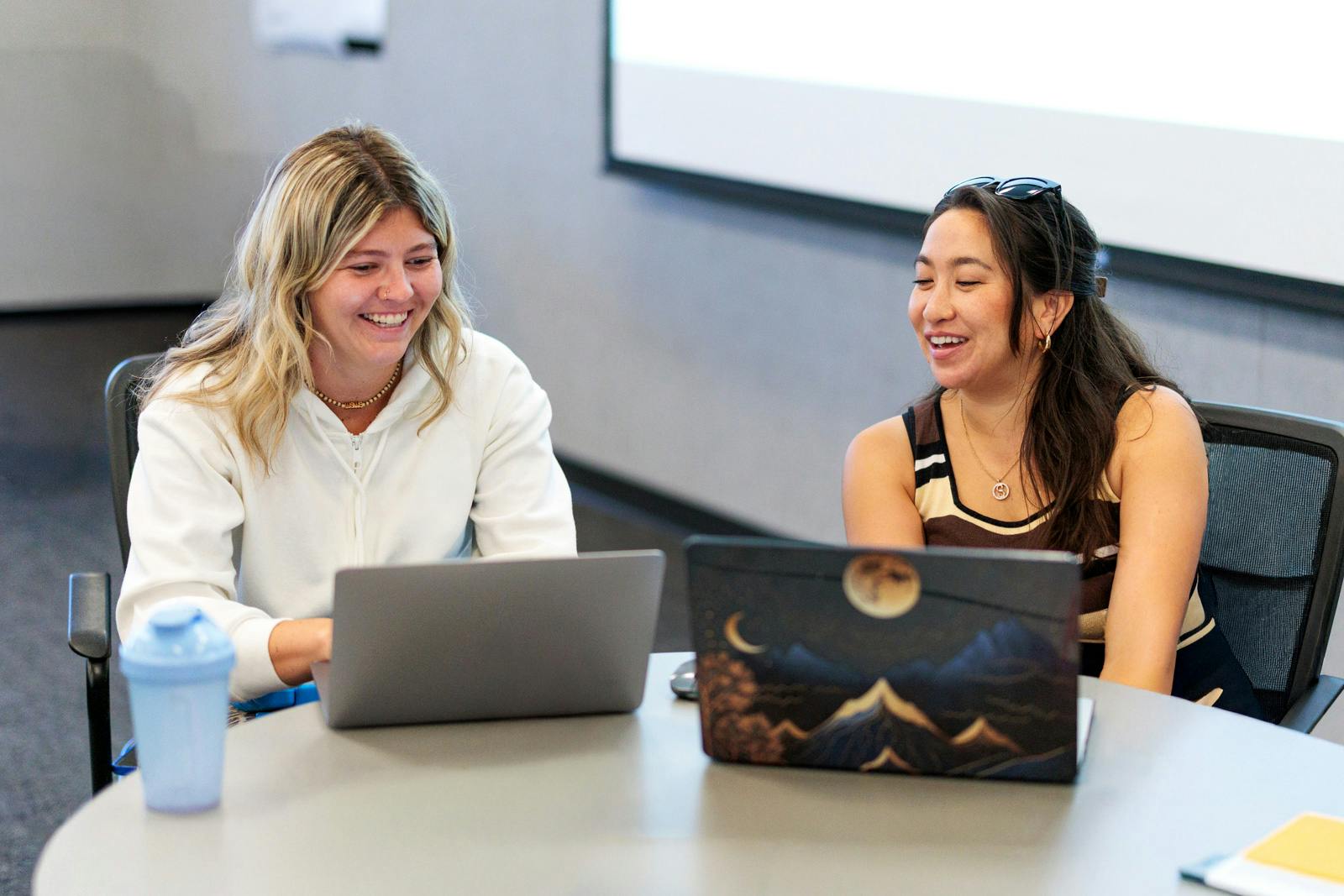 Two women smiling and working at laptops in a training room.