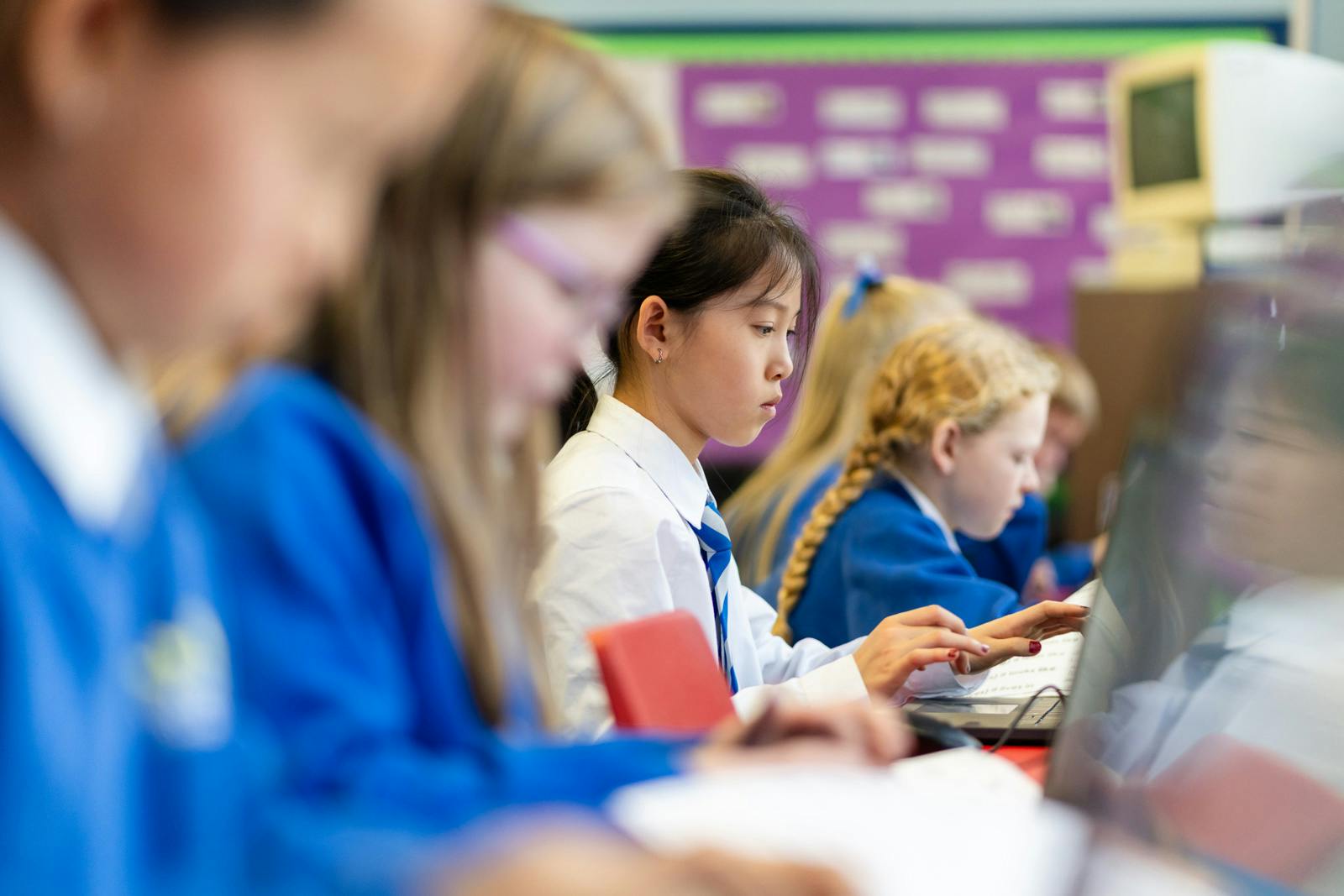 Students working at laptops in a classroom, with one girl in focus typing while others concentrate beside her.