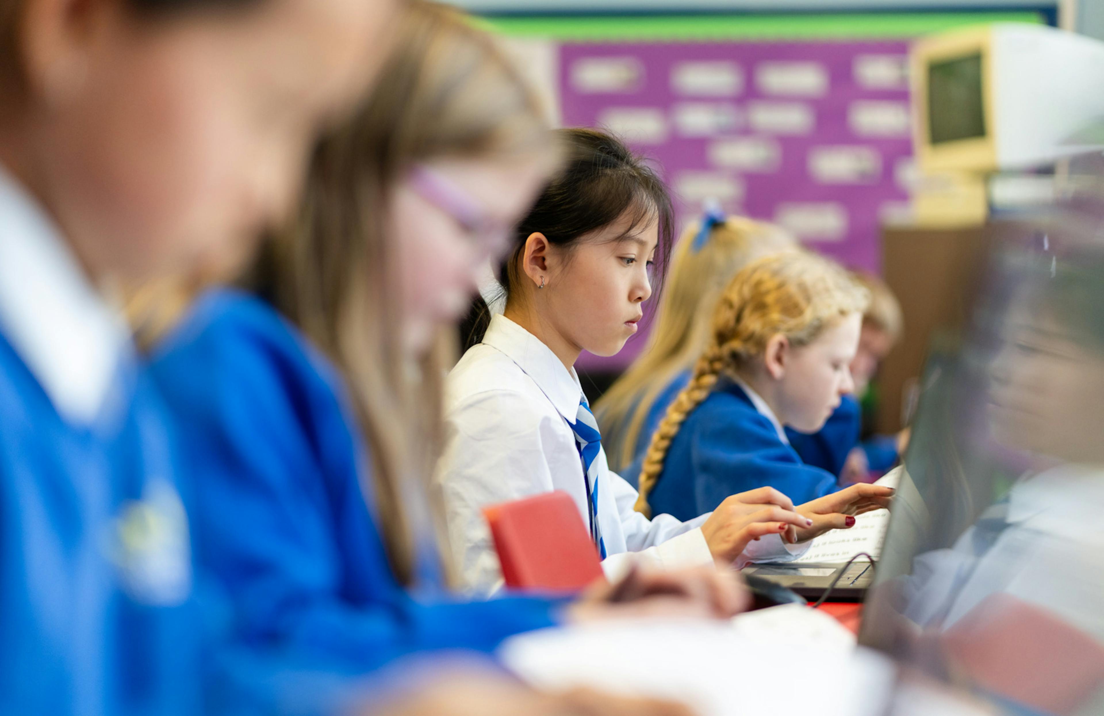 Students working at laptops in a classroom, with one girl in focus typing while others concentrate beside her.