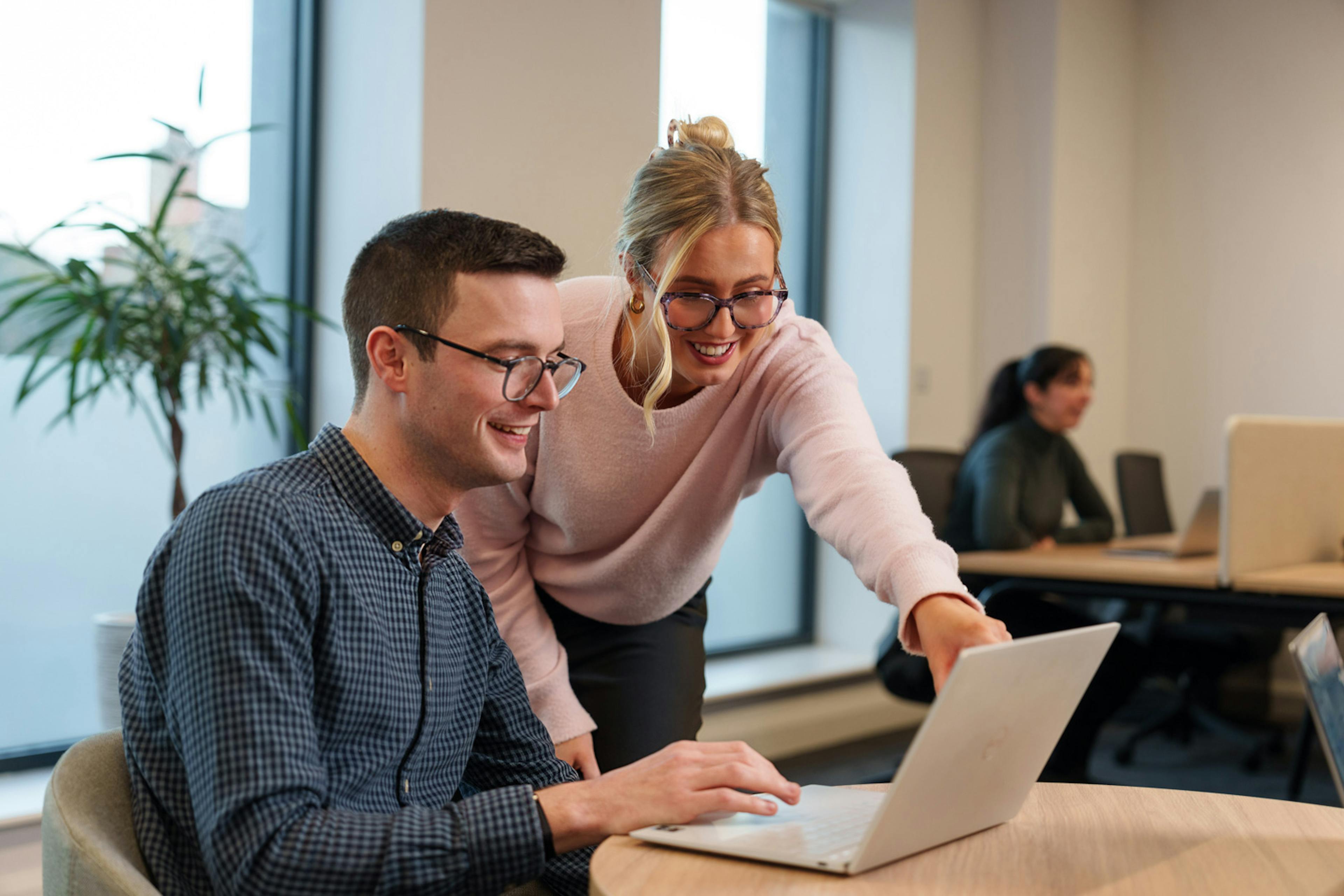 Two smiling colleagues in an office. One sitting at a computer, while the second stands beside them pointing at the computer screen.