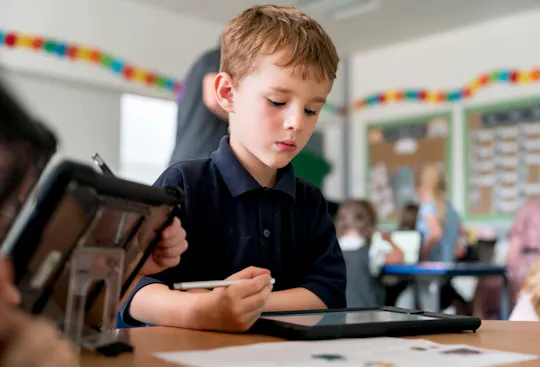Young student using a stylus on a tablet during class, focused on completing a digital assignment while classmates work in the background.