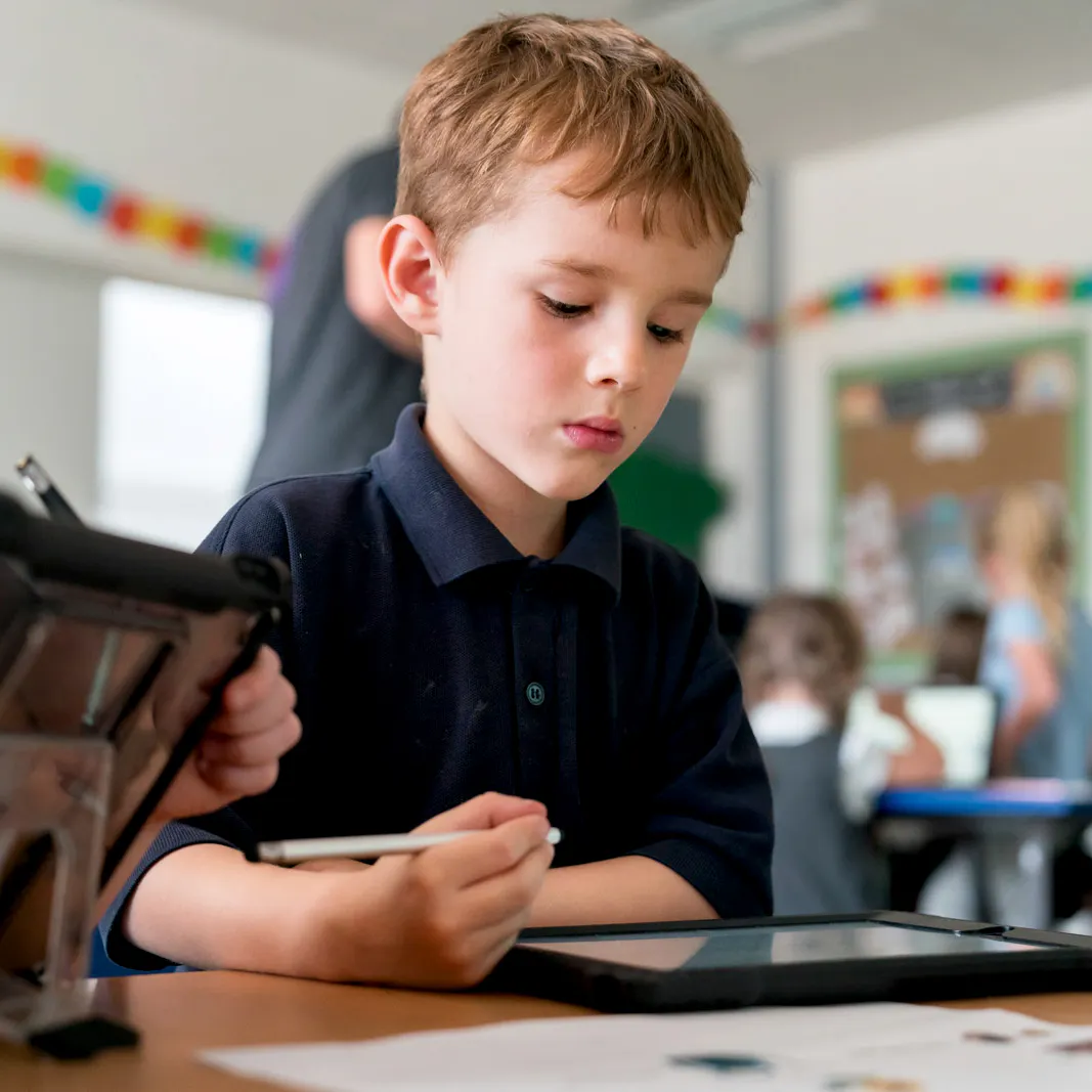 Young student using a stylus on a iPad during class, focused on completing a digital assignment while classmates work in the background.