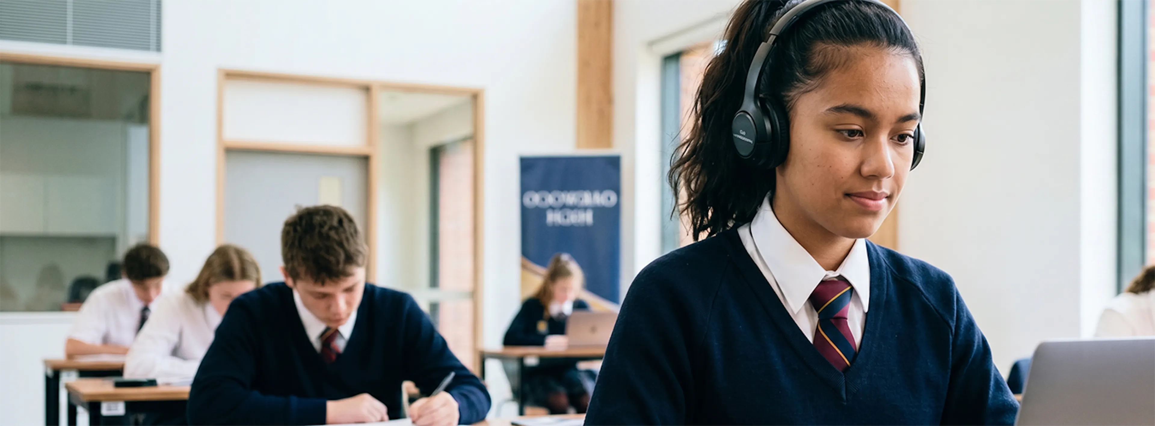 Student wearing headphones uses a laptop during a classroom exam while other students work at desks.
