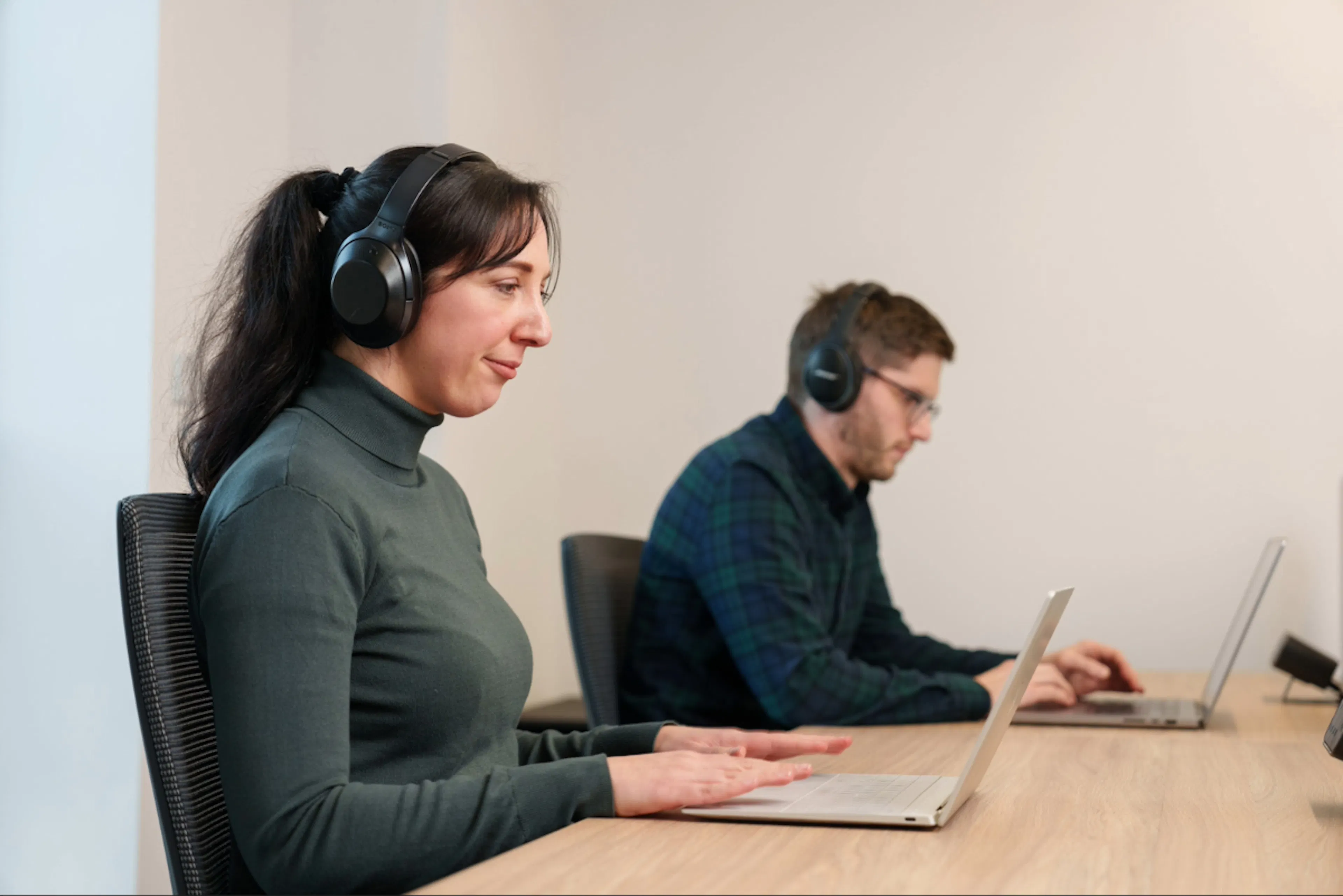 Two office employees sitting at a desk, both wearing wireless headphones and working on a computer