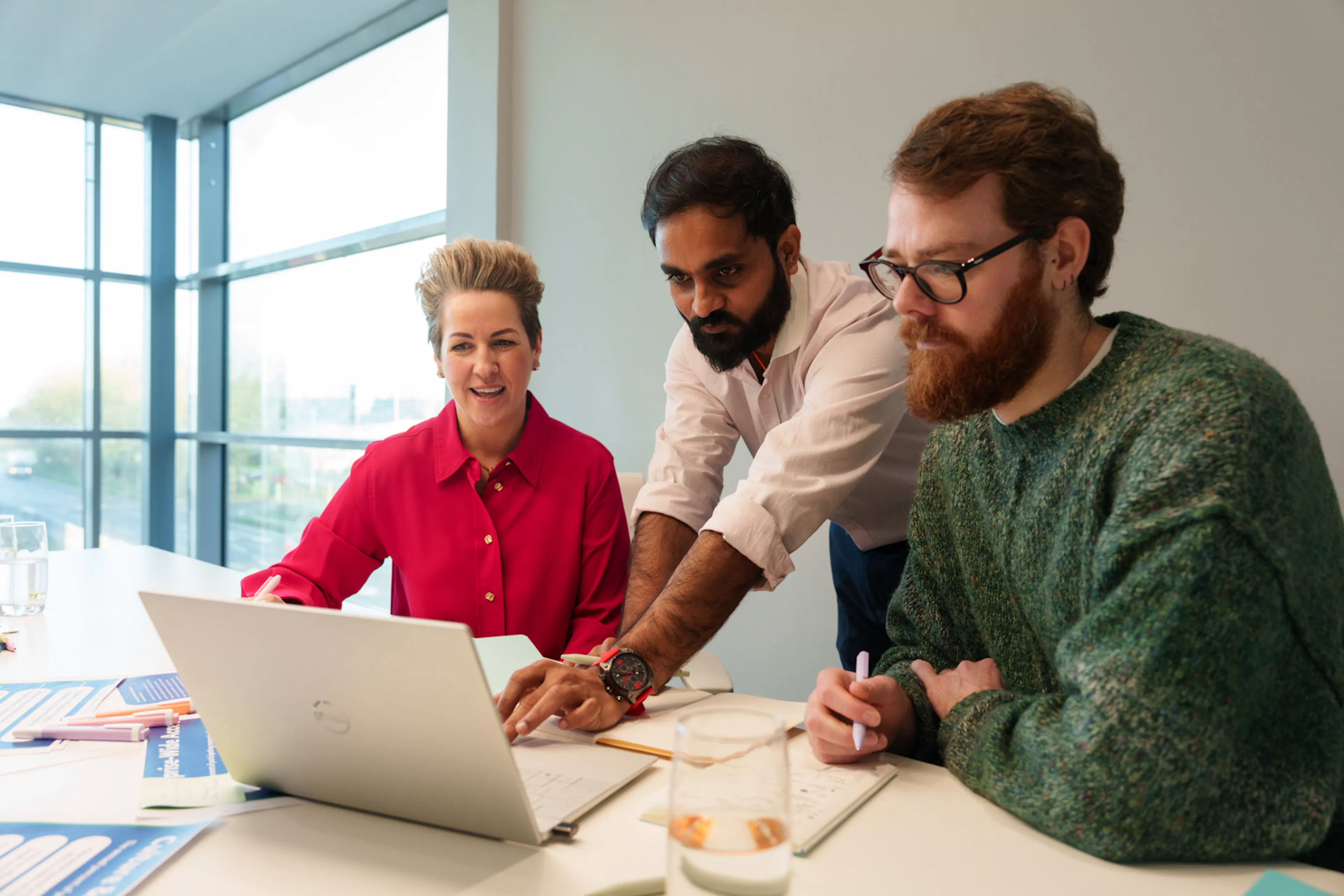 Three employees in a meeting setting. One employee is standing and pointing at a laptop screen