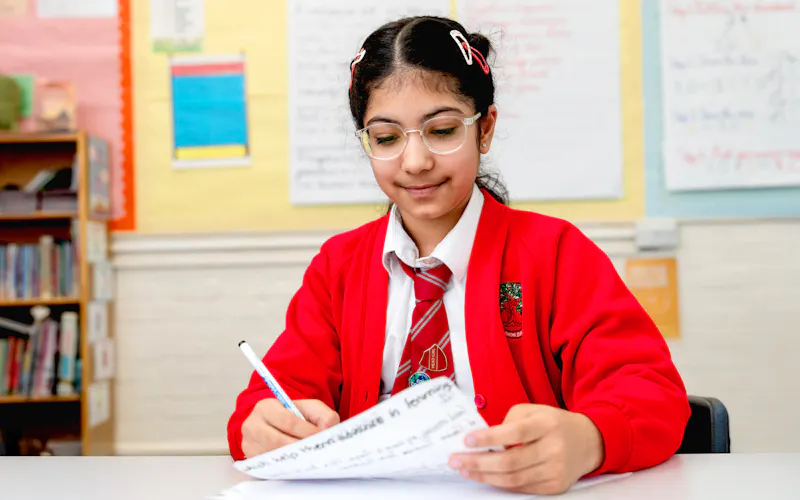 Student in a classroom writes on a worksheet, reviewing her work at a desk during a lesson.