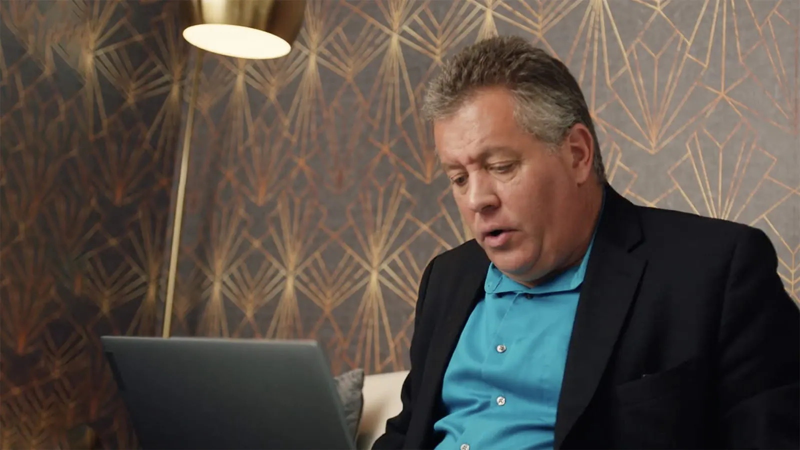A man (Peter) in a dark blazer and blue shirt sits working on a laptop in a warmly lit room with patterned wallpaper.