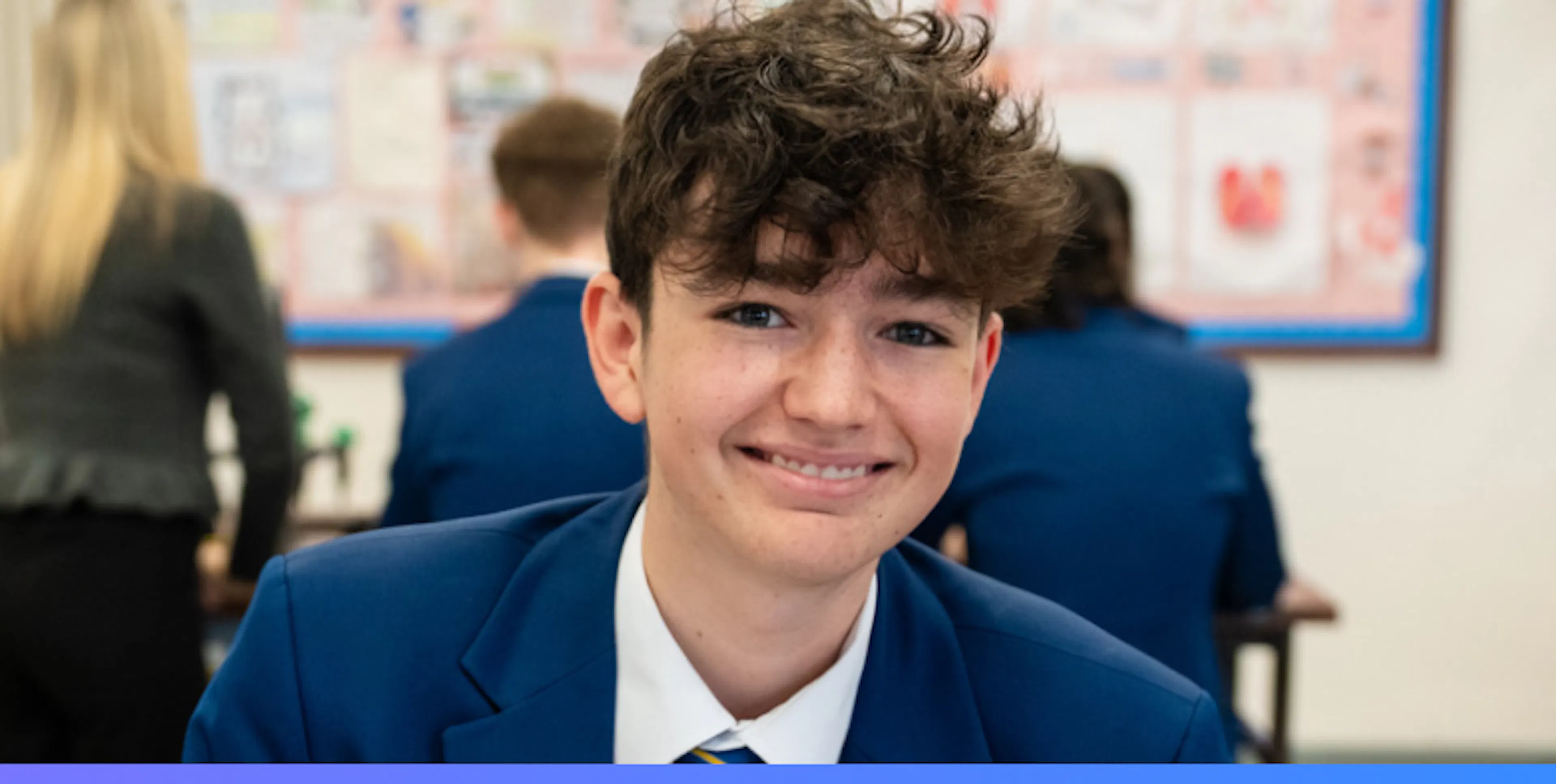 Smiling student in a blue school uniform looking at the camera while classmates work in the background.