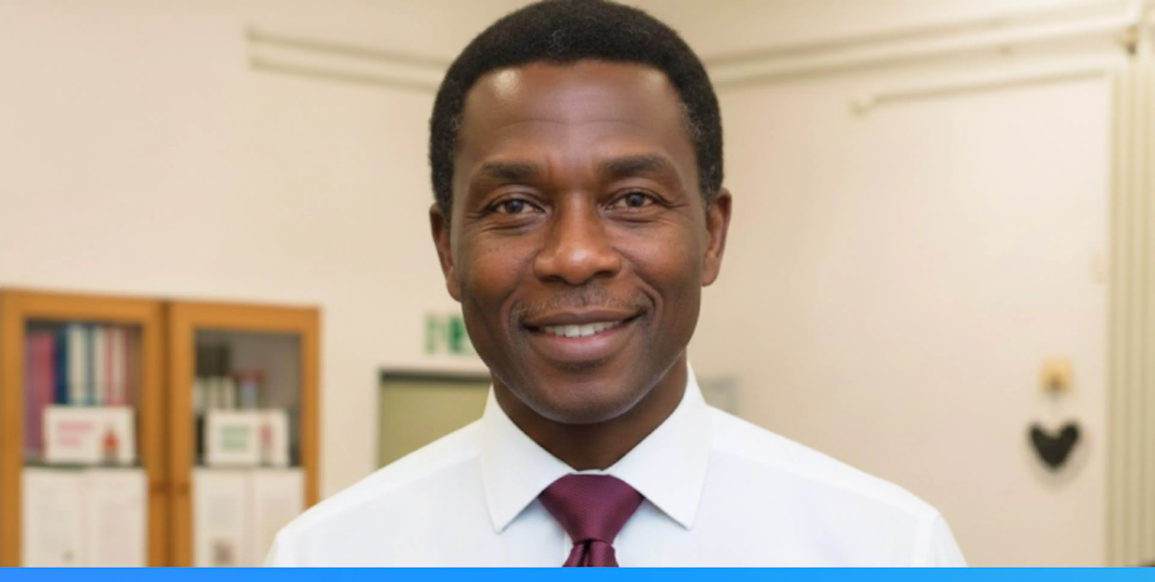 Teacher smiling, wearing a white shirt and burgundy tie in an school setting.