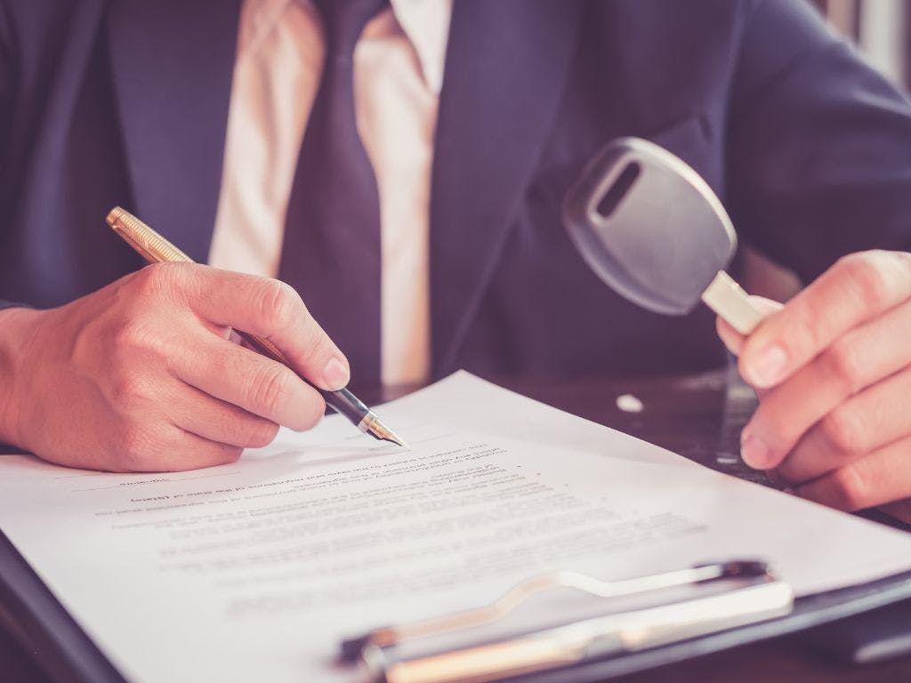 Jaki leasing na samochód ciężarowy wybrać? Porównanie dostępnych form finansowania; mężczyzna w garniturze podpisujący dokument i trzymający kluczyk do pojazdu w drugiej ręce