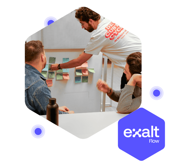 Tres personas discutiendo alrededor de una pizarra blanca llena de notas adhesivas, con el logo de eXalt Flow a su lado.