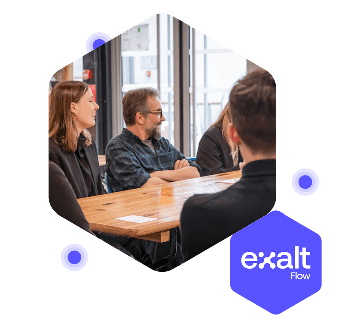 Varias personas sentadas alrededor de una mesa, asistiendo a una reunión, con el logo de eXalt Flow a su lado.