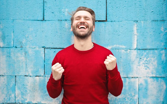 Man in red shirt expressing joy standing in front of blue wall.