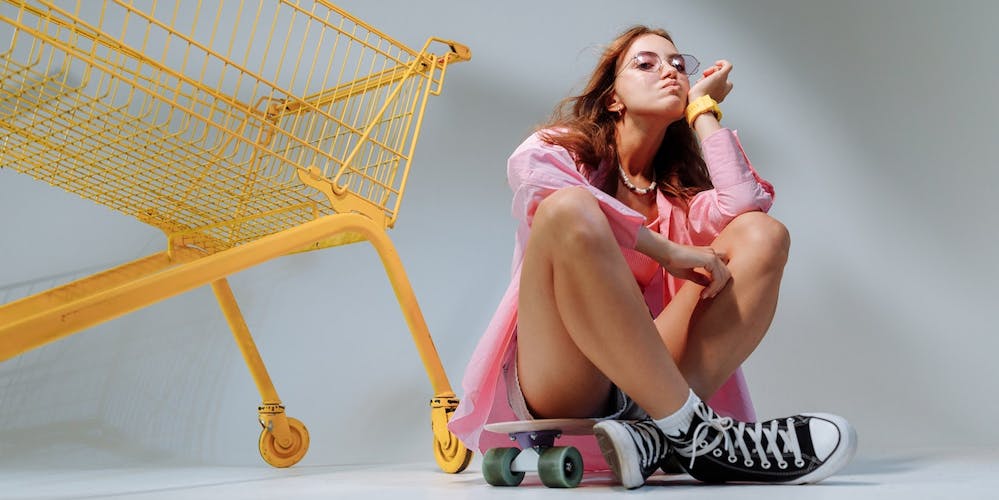 Frau sitzt auf Skateboard neben gelbem Einkaufswagen Frau sitzt auf Skateboard neben gelbem Einkaufswagen