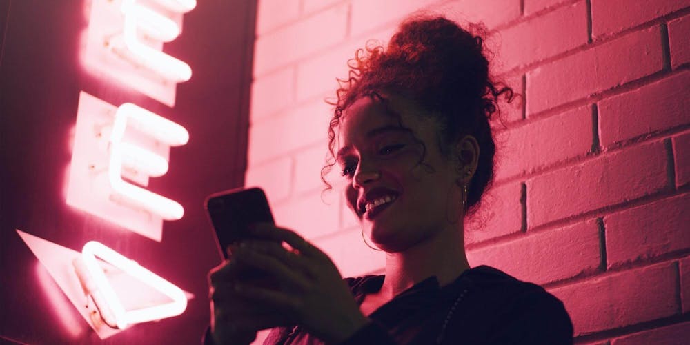 Frau steht vor einer Steinwand mit einem pinken Neonschild und scrollt am Smartphone Frau steht vor einer Steinwand mit einem pinken Neonschild und scrollt am Smartphone